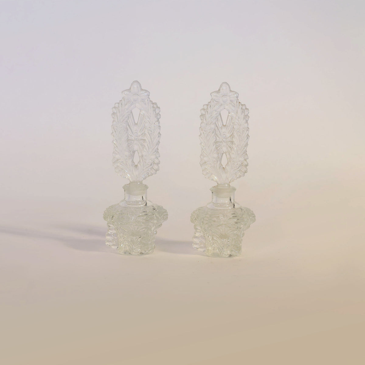 A pair of ornate, clear glass perfume bottles from Euro Trash Annie’s “Vintage Pressed Pair of Glass Perfume Bottles” collection stand side by side on a light, neutral background, their intricate matching stoppers capturing the timeless elegance of vintage vanity decor.