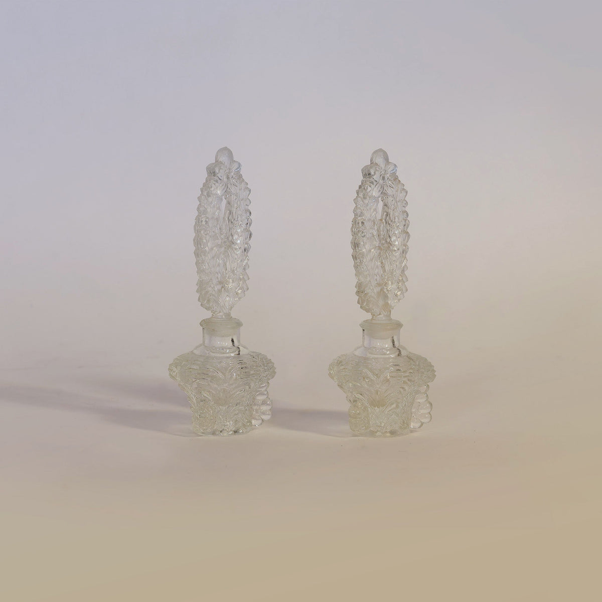 The Vintage Pressed Pair of Glass Perfume Bottles by Euro Trash Annie are displayed side by side on a neutral background, each adorned with matching tall, textured stoppers that cast soft shadows. These decorative bottles bring timeless elegance to any vintage glass decor collection.