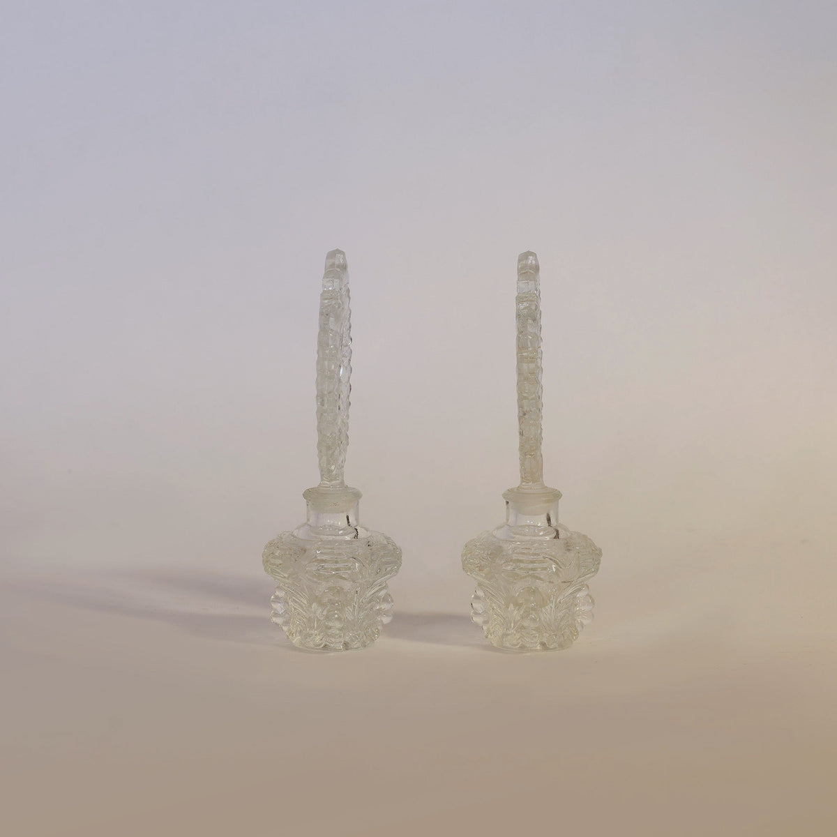 The Vintage Pressed Pair of Glass Perfume Bottles by Euro Trash Annie features two ornate bottles with long, decorative stoppers standing side by side on a neutral background. Their intricate pressed glass design reflects the light, showcasing delicate craftsmanship and timeless appeal as standout vintage glass decor pieces.