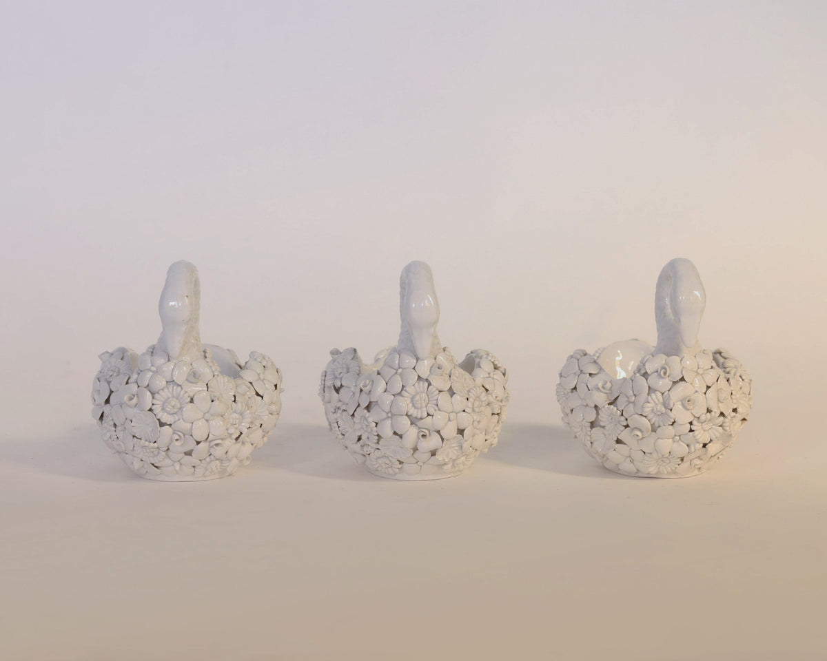 Three white ceramic planters shaped like swans, each adorned with delicate sculpted flowers and leaves, are arranged in a row against a plain, light background. The Vintage Set Of Floral Swan Planters by Euro Trash Annie features exquisite floral detailing, making them charming decorative accents for any elegant display.