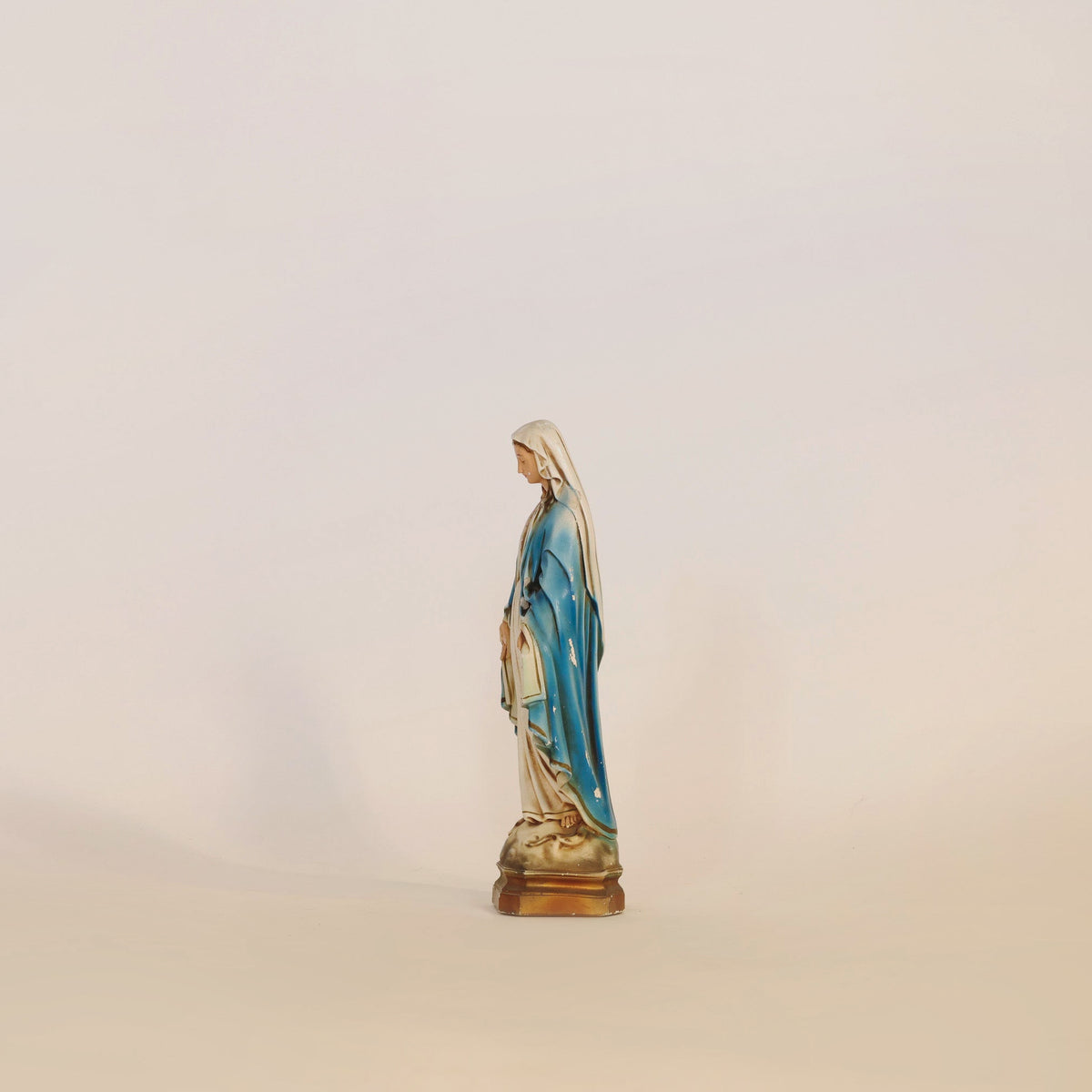 The Euro Trash Annie Vintage Virgin Mary Statue stands on a simple surface, adorned in a blue and white robe with hands joined in prayer against a beige backdrop. This graceful religious sculpture brings an element of spiritual home decor to any space.