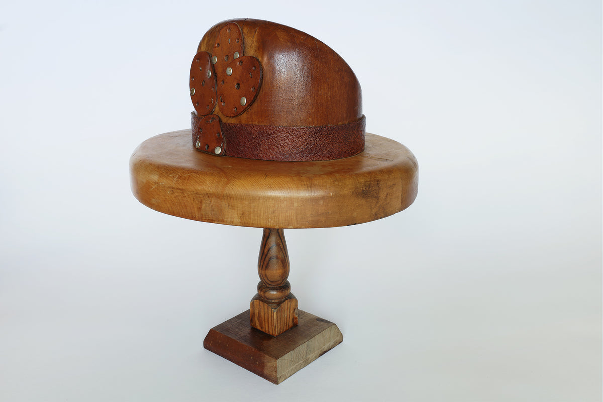 The Euro Trash Vintage Wooden Millinery Hat Mold, featuring a band and heart-shaped decorations, is displayed on a wooden pedestal against a plain white background.