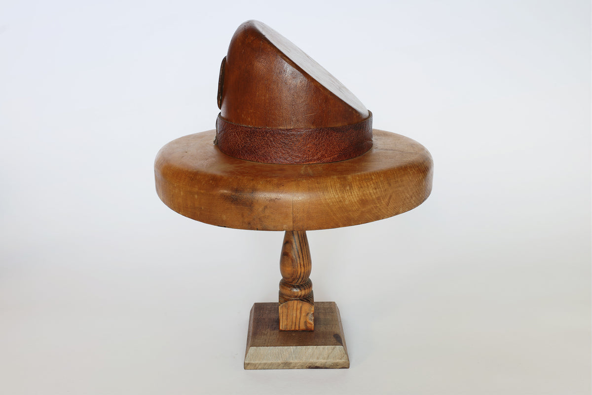 The Euro Trash Vintage Wooden Millinery Hat Mold is a sculpted wooden piece on a stand, featuring a rounded brim, angled crown, and textured band at the base. Displayed against a plain white background, this distinctive object functions both as an elegant tool for millinery work and as a striking decorative accent.