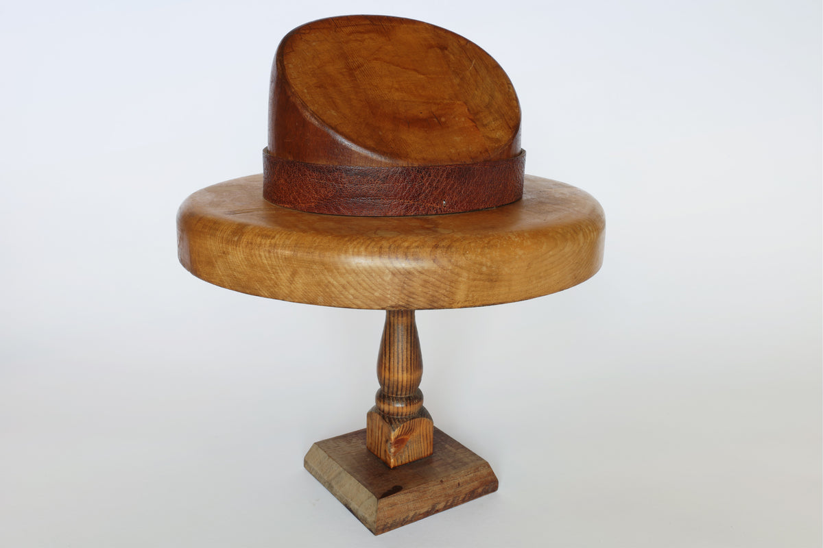 The Euro Trash Vintage Wooden Millinery Hat Mold, showcasing a wide brim and domed crown with a dark leather band, is presented on a turned wooden pedestal stand against a white background. This collectible piece serves as both a functional millinery tool and an elegant decorative object.