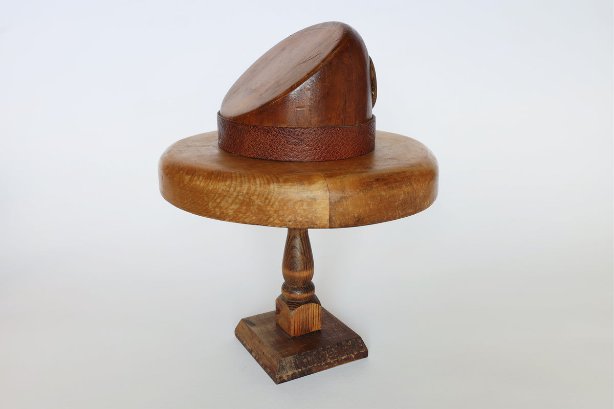The Euro Trash Vintage Wooden Millinery Hat Mold, shaped like a head and set on a turned wooden stand, features a round, brown textured band resembling a hat brim. This piece serves as both a functional millinery tool and an eye-catching decorative accent.