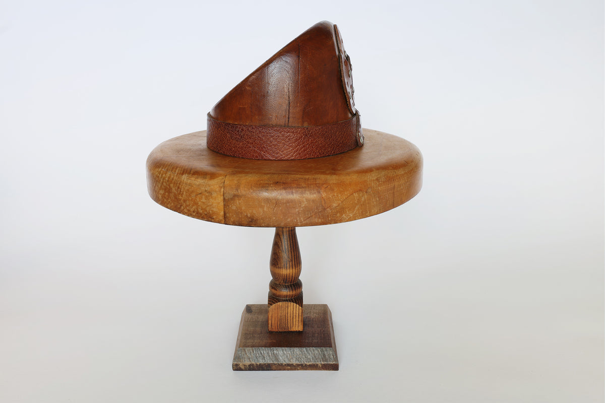 A Euro Trash Vintage Wooden Millinery Hat Mold, featuring a round display form with a single pedestal and square base, is shown holding a brown leather hat with a textured band against a plain white background.