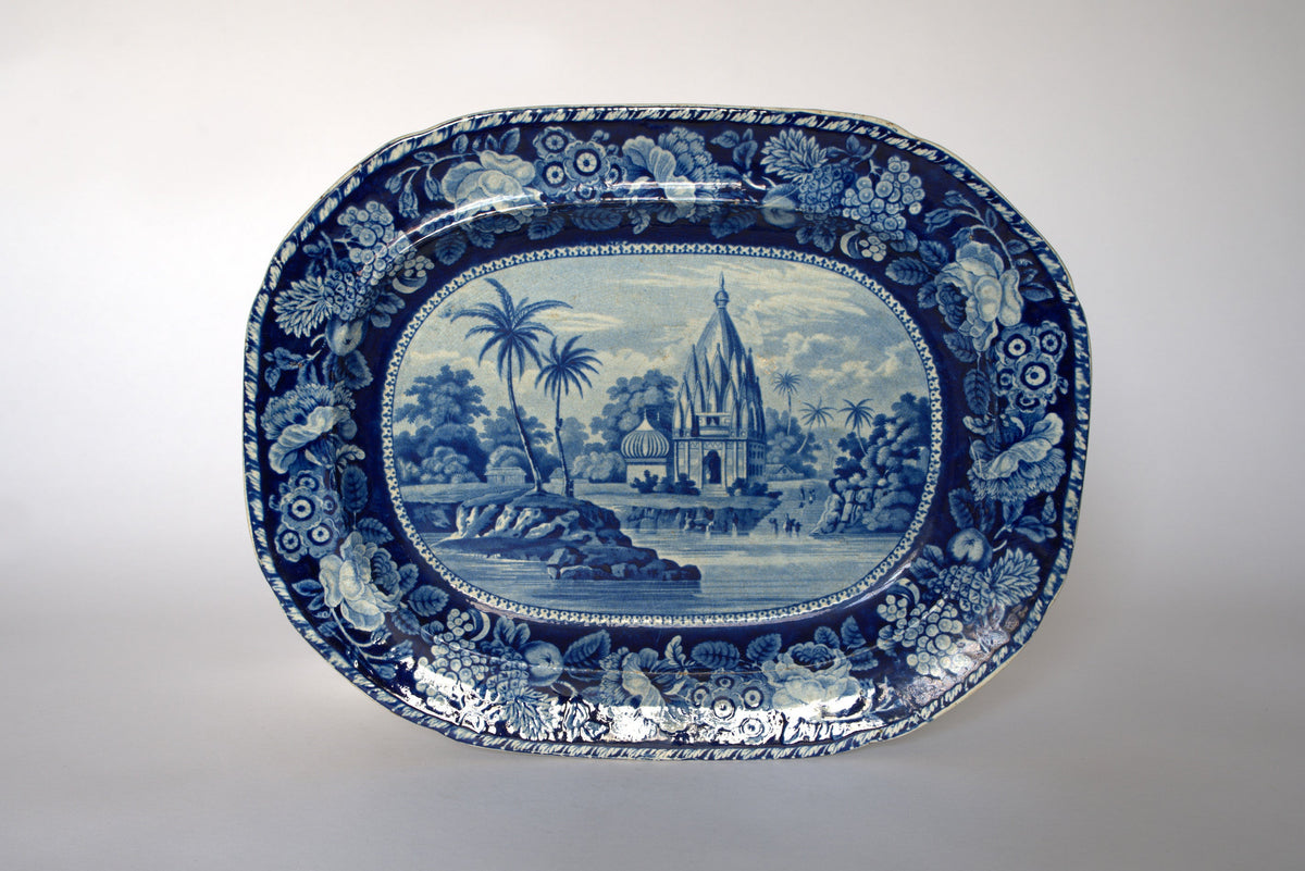 The Euro Trash Vintage Blue & White Floral Transferware Serving Platter features a central temple scene by a river with trees and people, framed by a floral border—ideal as a unique vintage serving piece or decorative accent.