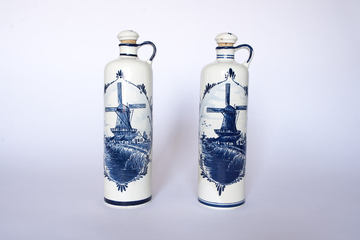 The Euro Trash Vintage Blue & White Windmill Motif Ceramic Bottle Decanters set features two tall handled bottles with classic blue windmill and landscape designs, displayed against a plain white background.