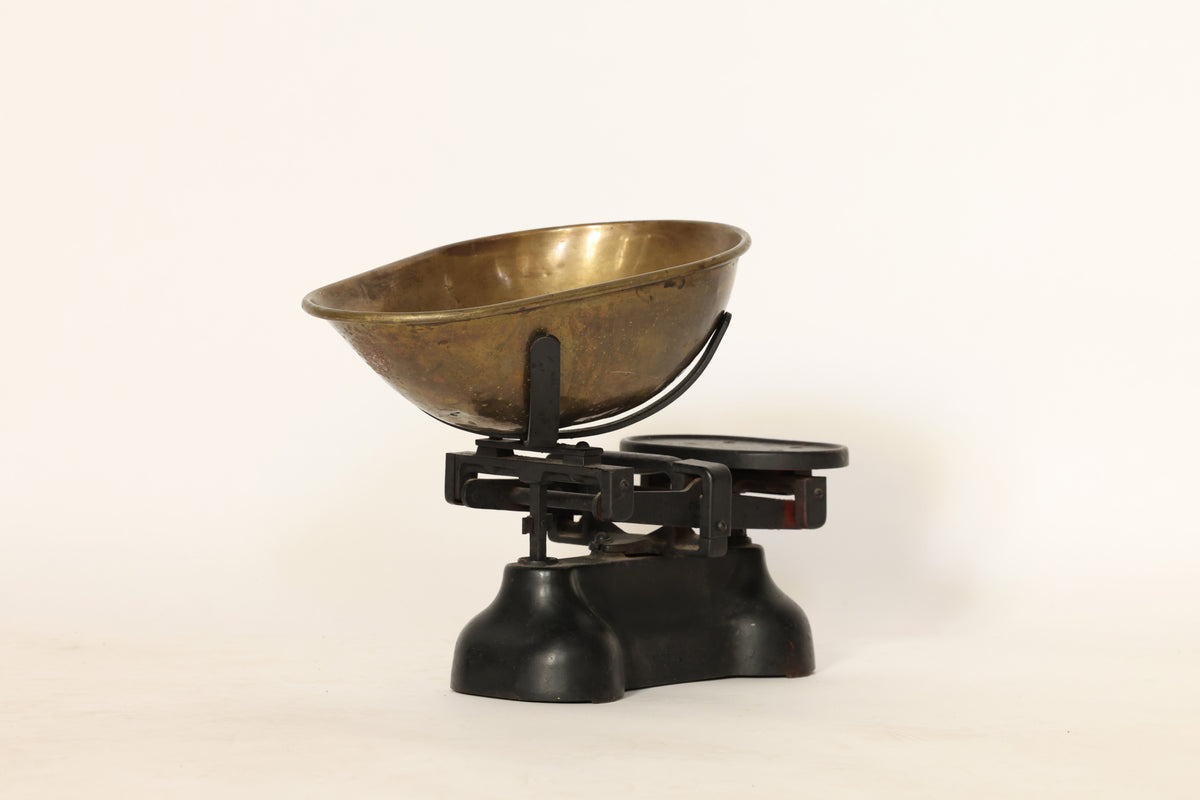 The Vintage Brass Balance Scale by Euro Trash Annie features a large brass bowl on one side and a flat metal platform on the other, all set on a black metal base against a plain white background.