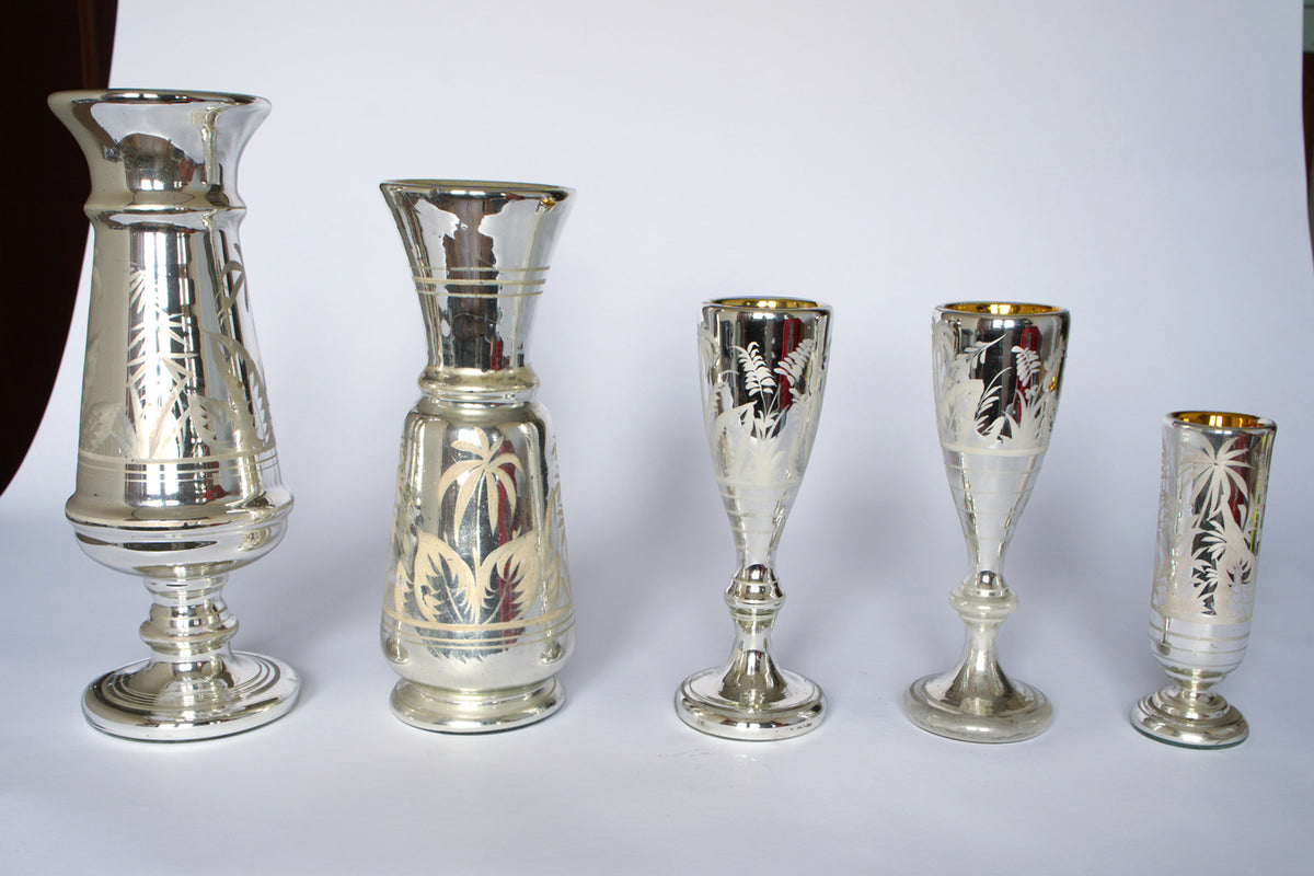 The Euro Trash Vintage Etched Mercury Glass Vessel Set features five silver vases and goblets of varying heights, each with botanical motifs and a reflective finish, arranged in a row on a white background.
