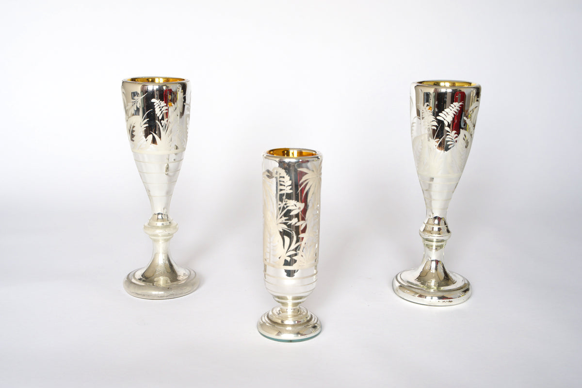 The Vintage Etched Mercury Glass Vessel Set by Euro Trash features three silvered vessels with frosted botanical patterns and gold-toned interiors. Two taller pieces frame the shorter center one, shown on a white background.