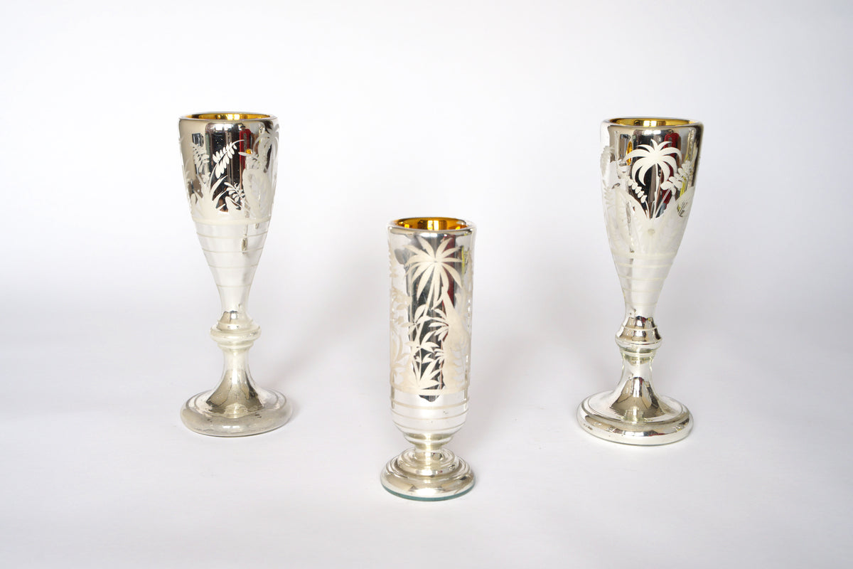 The Vintage Etched Mercury Glass Vessel Set by Euro Trash features three ornate silvered vessels with engraved palm tree motifs on a white background. The central piece is shorter and cylindrical, flanked by two taller, wider-based vessels.