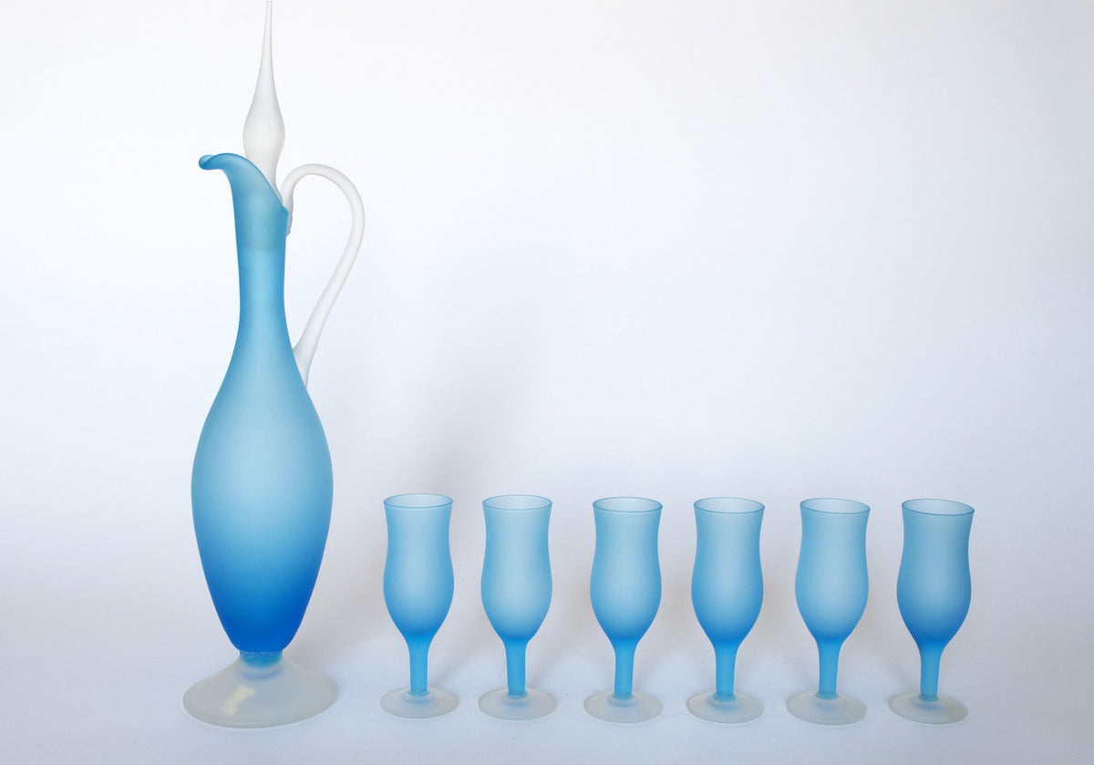 The Euro Trash Vintage Frosted Blue Glass Decanter and Stemware Set features a tall blue decanter with white handle and stopper, paired with five matching blue stemmed glasses, all displayed in a row on a white background.
