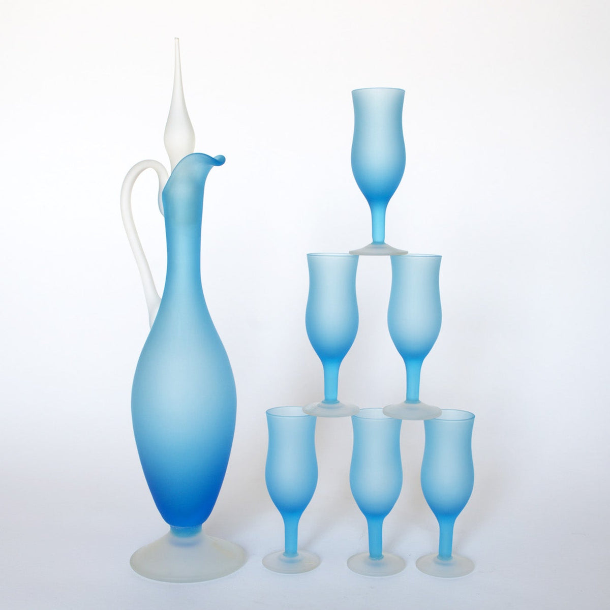 The Euro Trash Vintage Frosted Blue Glass Decanter and Stemware Set features a tall blue decanter with a white handle and stopper beside six matching blue stemmed glasses, all displayed on a plain white background.