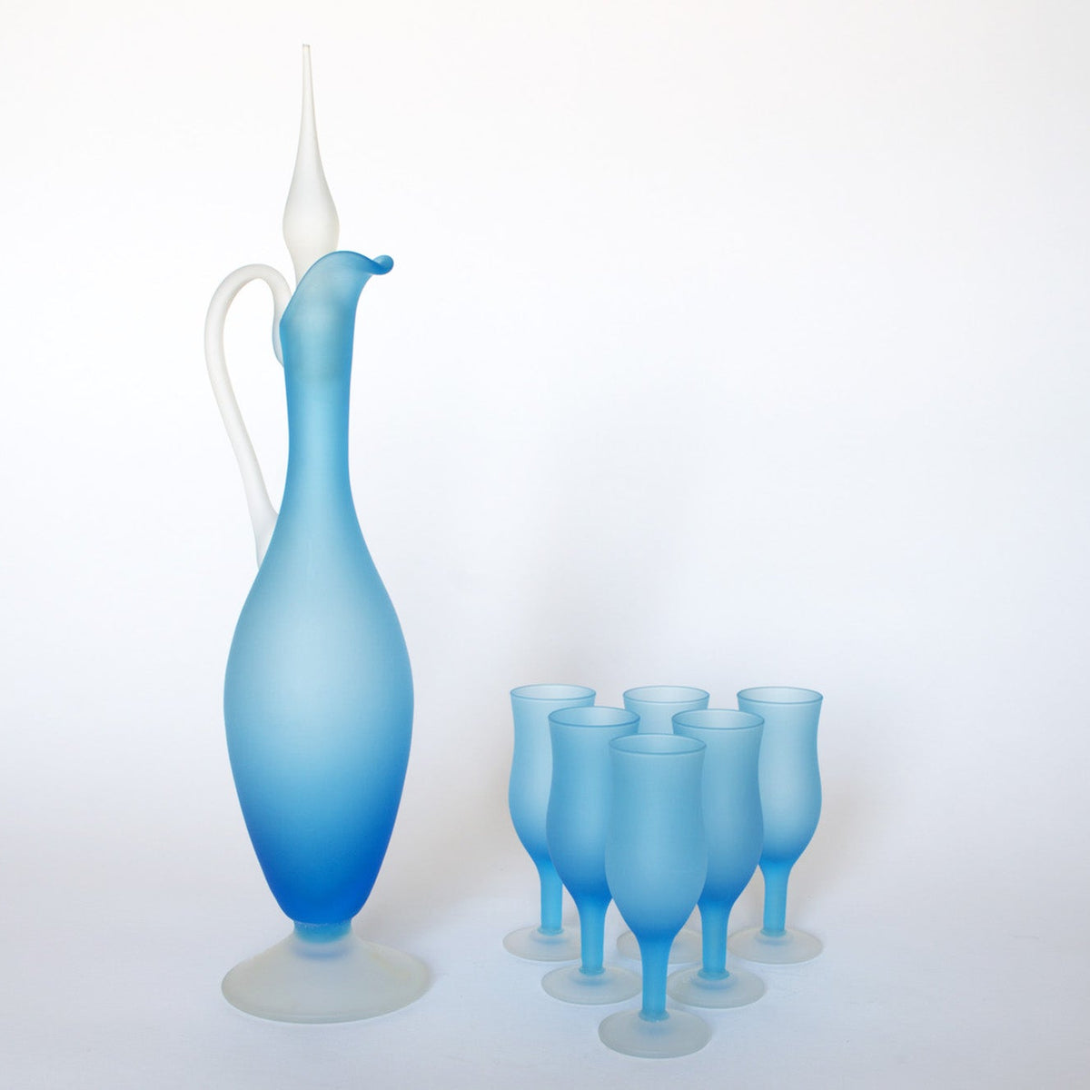 The Euro Trash Vintage Frosted Blue Glass Decanter and Stemware Set features a tall decanter with handle and stopper beside six elegant blue stemmed glasses, displayed on a plain white background.