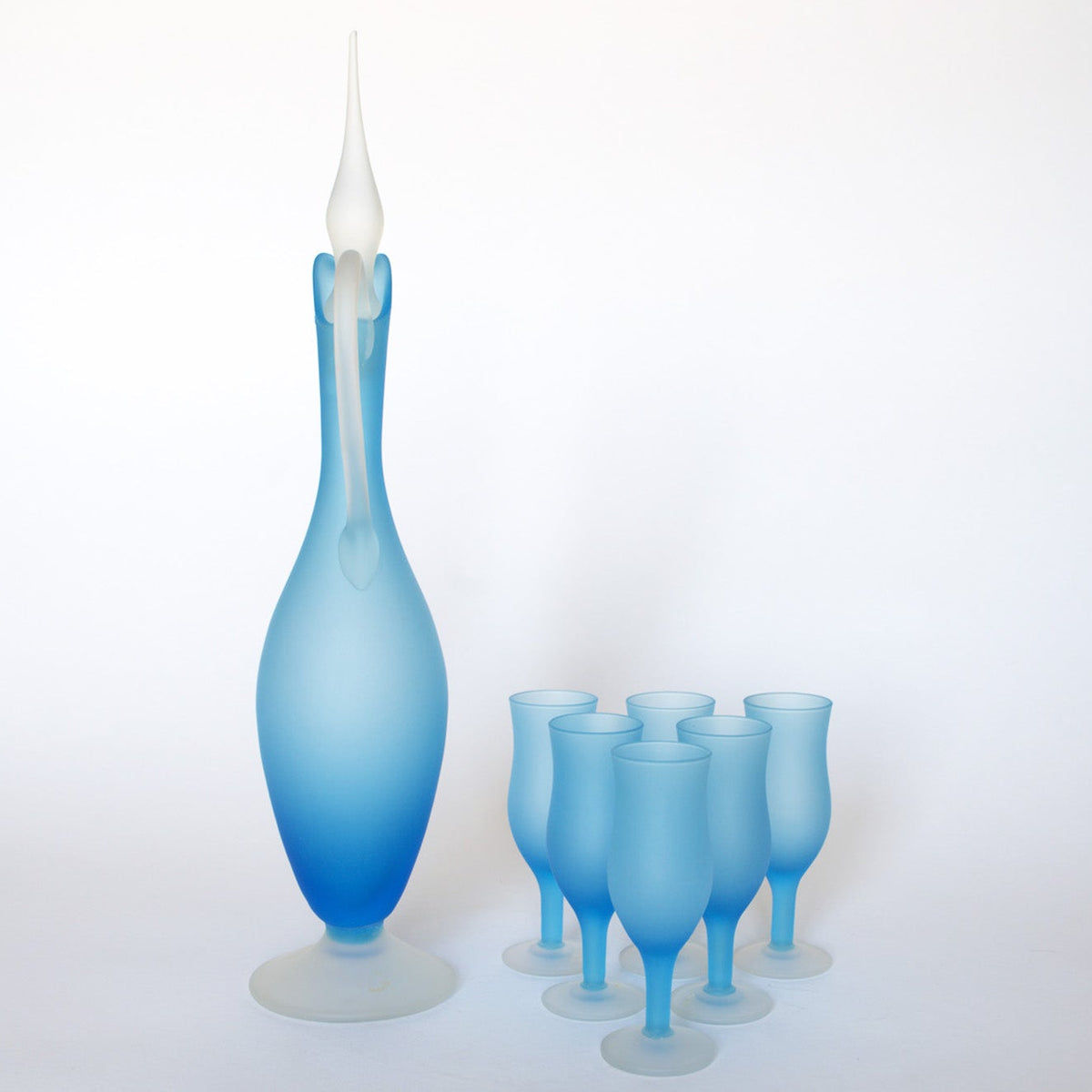 The Euro Trash Vintage Frosted Blue Glass Decanter and Stemware Set features a tall decanter with a pointed stopper alongside six matching blue stemmed glasses, all displayed on a white background.