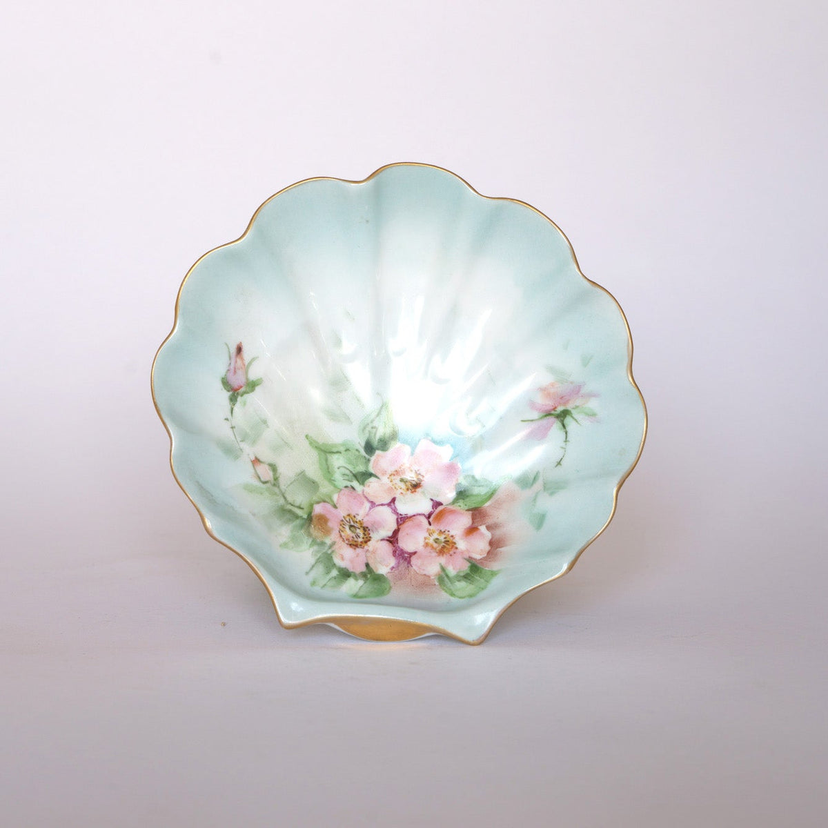The Euro Trash Vintage Hand-Painted Floral Porcelain Plate features pink flowers and green leaves on a light blue scalloped plate, accented with a gold rim along the wavy edge.