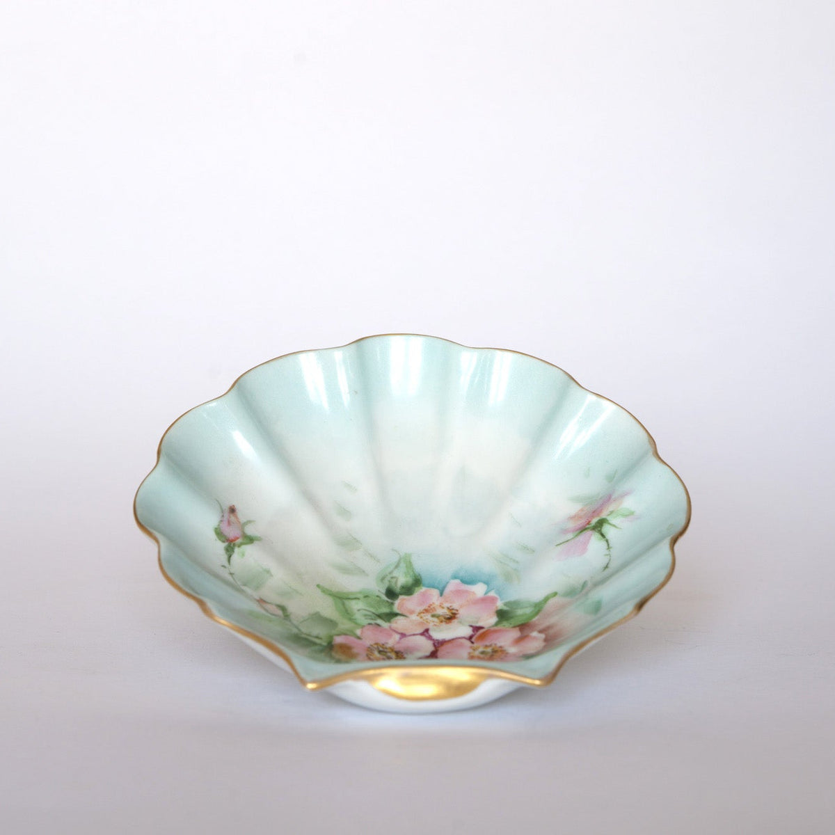 The Euro Trash Vintage Hand-Painted Floral Porcelain Plate features a scallop shape, gold rim, pink flowers, green leaves, and a pale blue-green glaze on a plain white background.