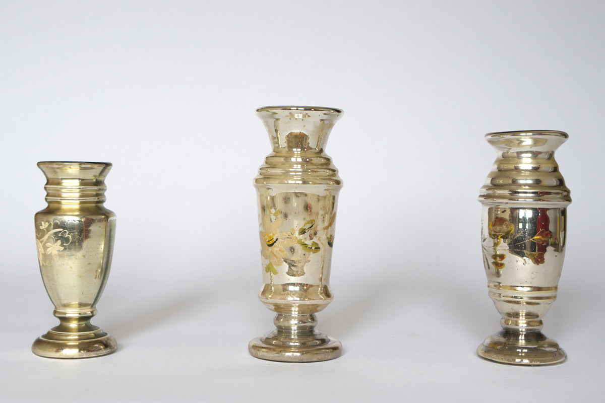 The Euro Trash Vintage Hand-Painted Mercury Glass Vase Set features three vases of different shapes and heights, each with a reflective silver finish and faded floral designs, displayed against a plain white background.