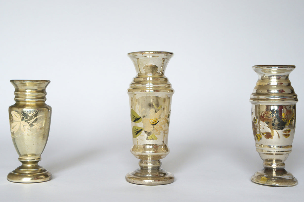 The Euro Trash Vintage Hand-Painted Mercury Glass Vase Set features three silvered vases with faded floral patterns, each uniquely shaped and showing charming signs of age, displayed against a plain white background.