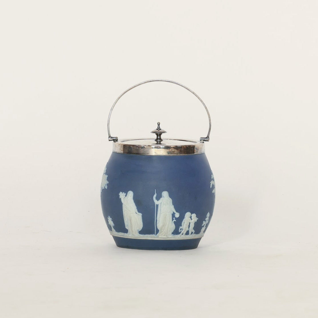 Introducing the Vintage Jasperware Wedgwood Style Biscuit Jar by Euro Trash Annie—a blue ceramic jar adorned with elegant white neoclassical relief designs, featuring a silver metal lid and a curved handle. Perfect as a decorative storage piece, this classic accessory is set against a plain white background.