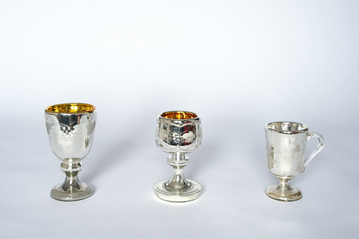 The Euro Trash Vintage Mercury Glass Stemmed Cup Set features three cups on a white background. Two have gold interiors, wide stems, and etched details; the third is smaller, with a handle and a plain silver interior.