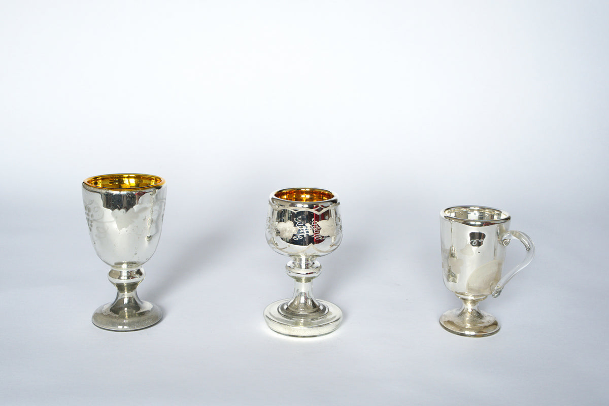 The Euro Trash Vintage Mercury Glass Stemmed Cup Set features three silver cups with gold interiors in distinct shapes and bases, including one with a handle, all displayed side by side on a plain white background.