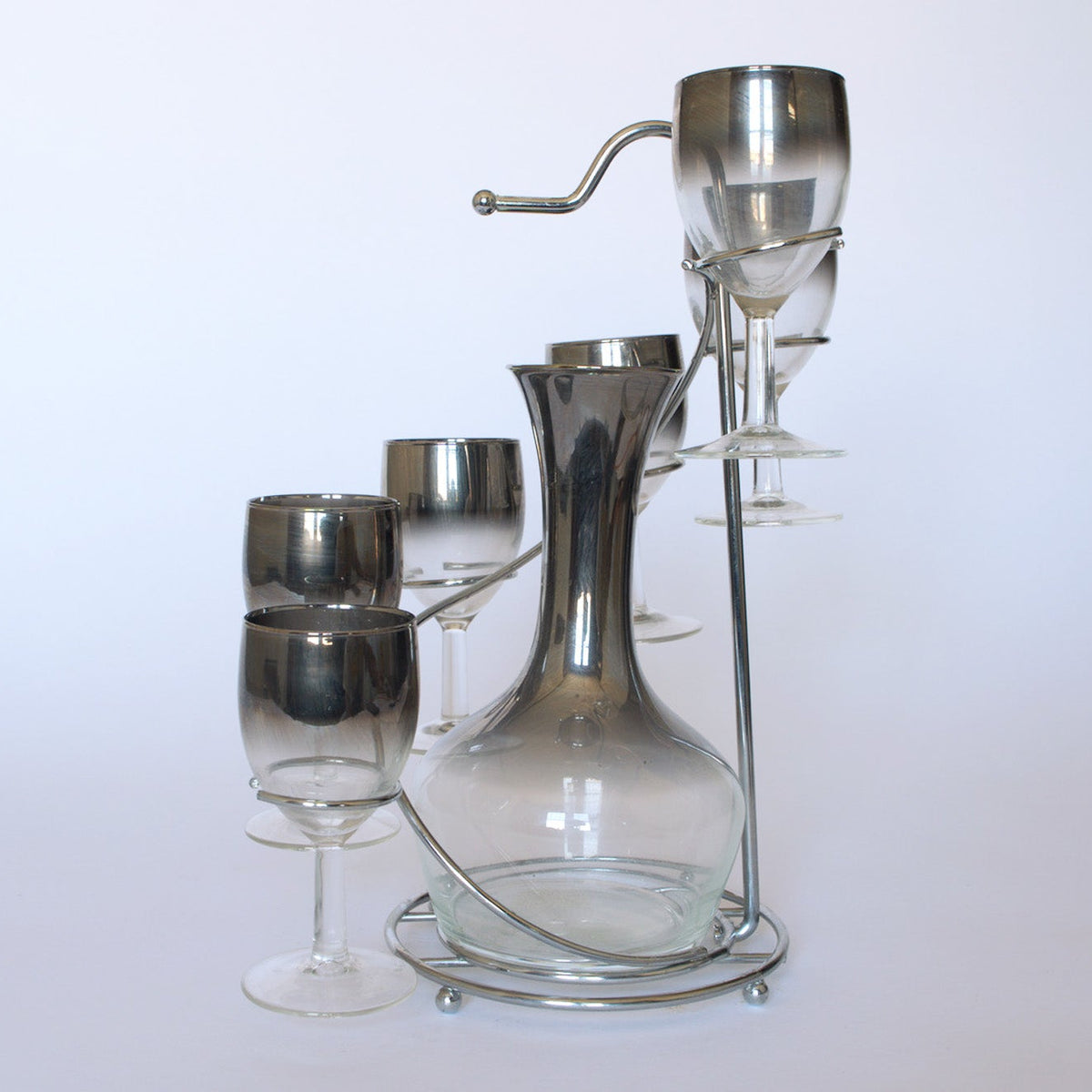 The Euro Trash Vintage Mid-Century Bar Set with Decanter includes a clear decanter in a metal stand and six smoked glass wine glasses, three hung upside down, blending modern minimalist and mid-century bar style.