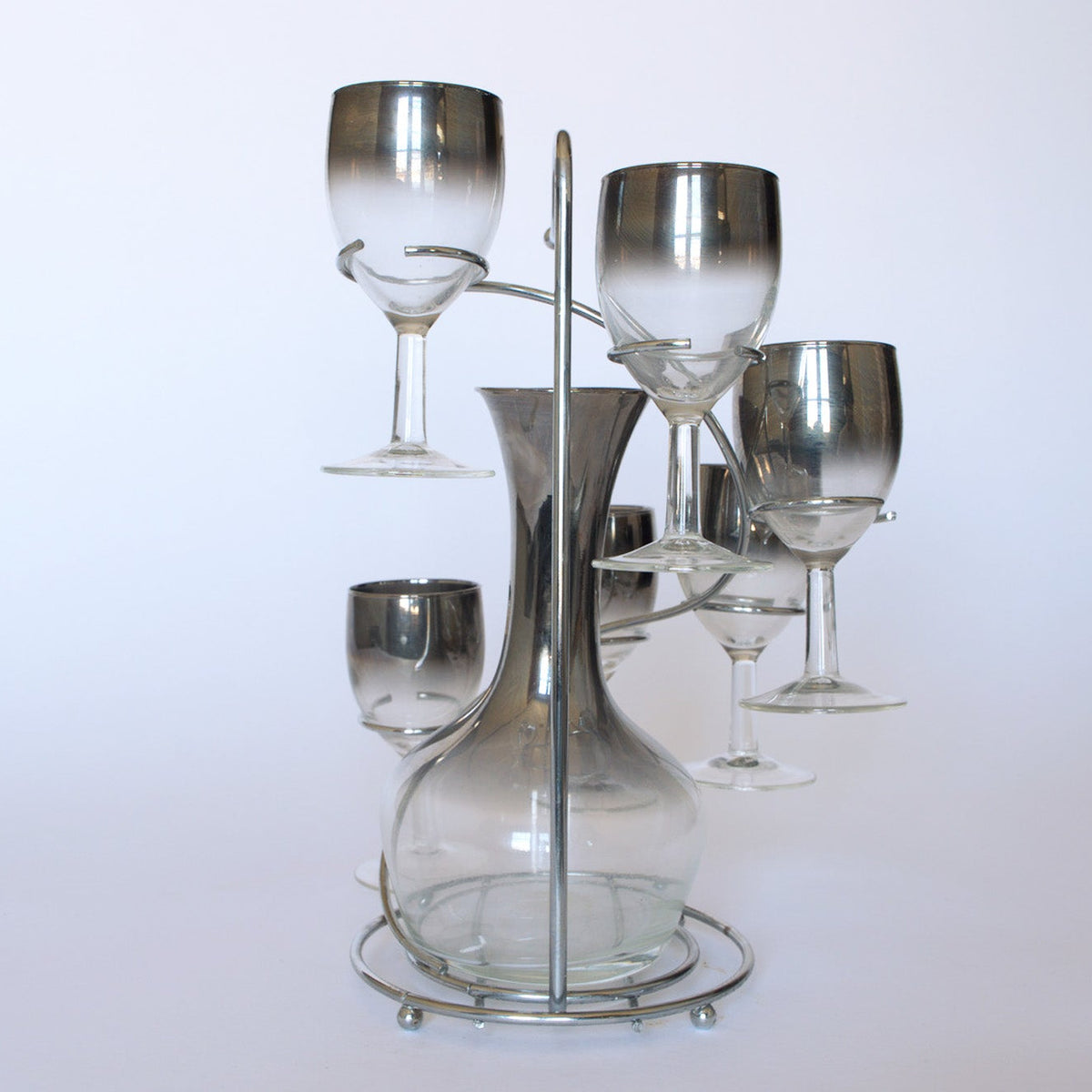 The Euro Trash Vintage Mid-Century Bar Set with Decanter features a clear decanter and five smoked glass wine glasses with silver rims, all set on a metal stand and shown against a white background.