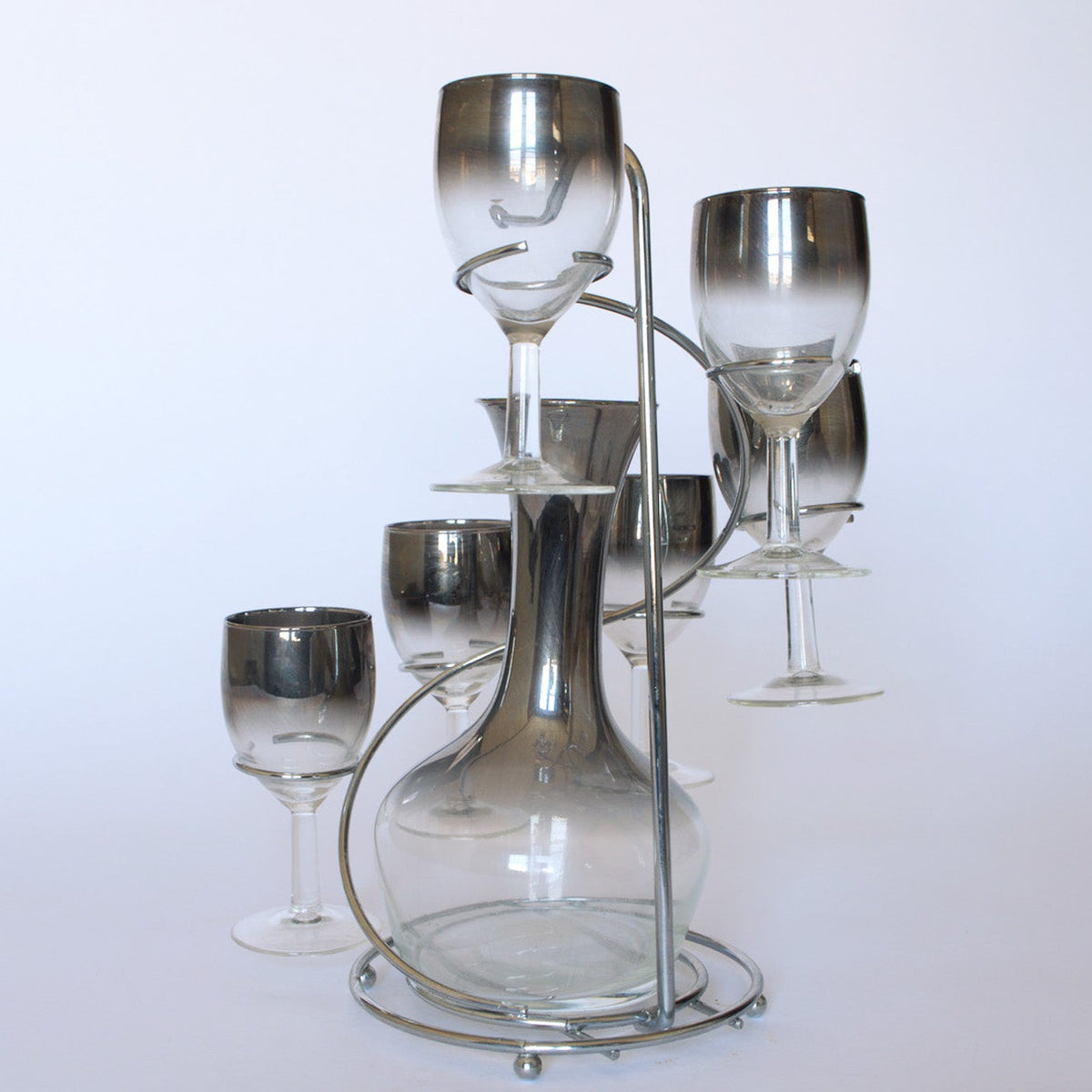 The Euro Trash Vintage Mid-Century Bar Set with Decanter features a clear decanter in a metal stand, surrounded by six smoked glass stemware pieces arranged in a circle against a plain white background.