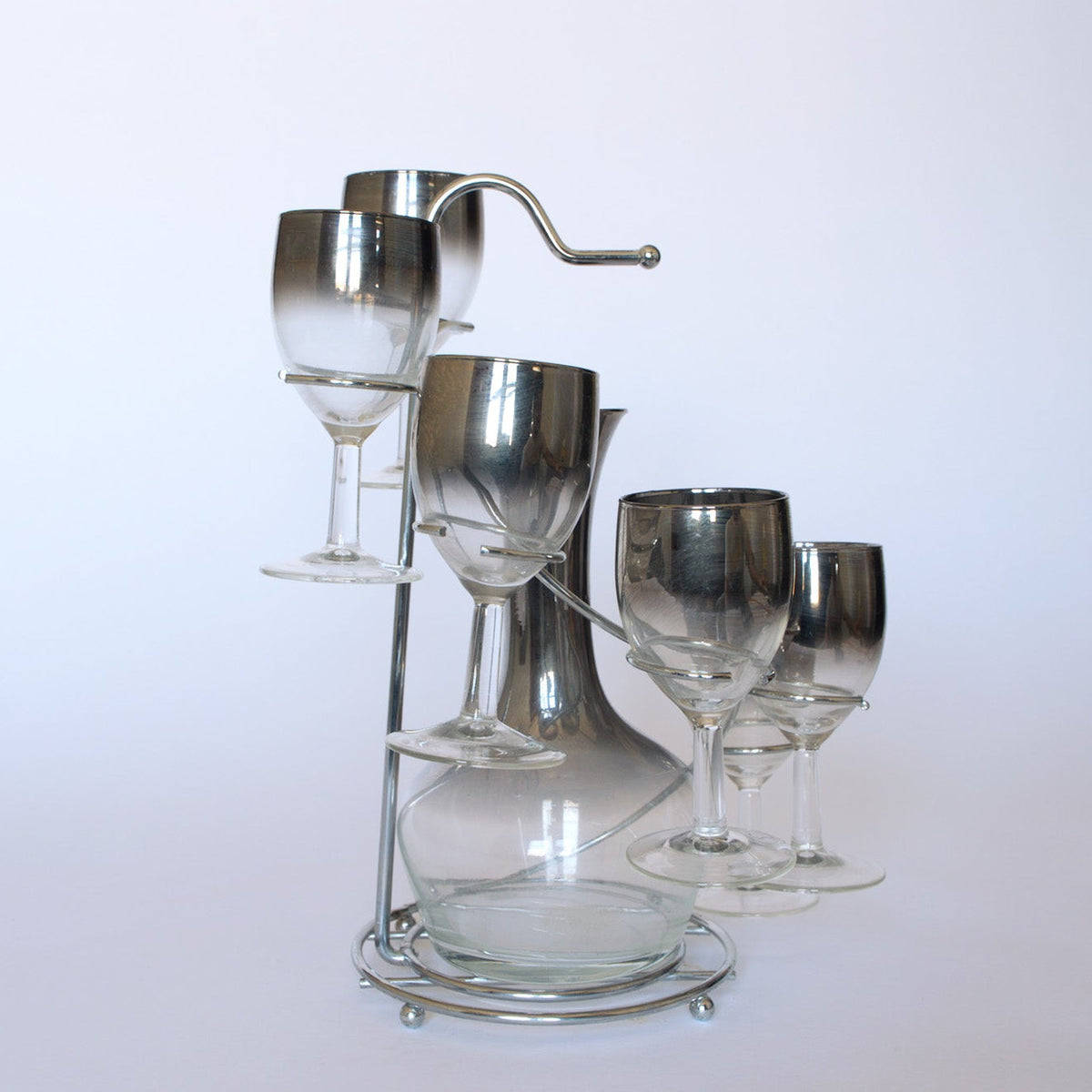 The Euro Trash Vintage Mid-Century Bar Set with Decanter features a metal-handled decanter and smoked glass stemware arranged on a sleek circular metal stand, shown against a plain white background.
