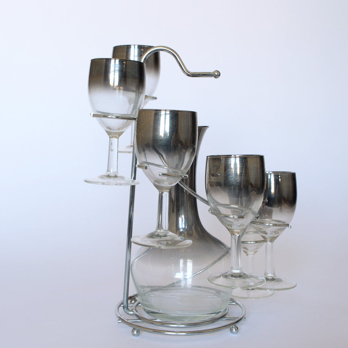 The Euro Trash Vintage Mid-Century Bar Set with Decanter includes a clear glass decanter and four wine glasses with silver rims on a sleek metal stand—perfect for adding elegance to any occasion.