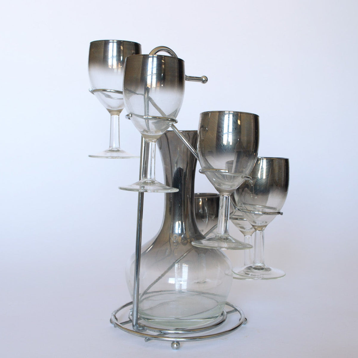 The Euro Trash Vintage Mid-Century Bar Set features a clear decanter with a metal handle, four matching wine glasses with metallic rims, and a circular stand, all shown against a plain white background.