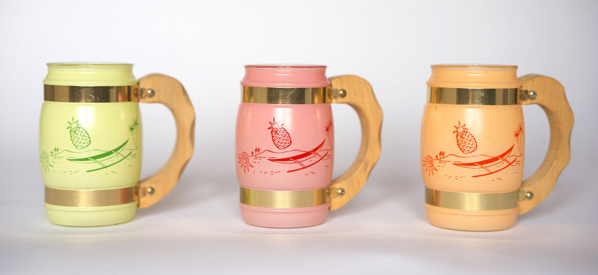 The Euro Trash Vintage Pastel Glass Mug Set features three mugs in green, pink, and orange with wooden handles and gold bands. Each displays a lounging pineapple in a hammock, shown side by side on a white background.