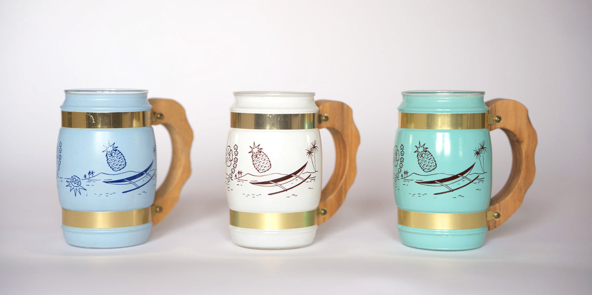 The Euro Trash Vintage Pastel Glass Mug Set includes three mugs with wooden handles and gold bands in light blue, white, and aqua, each featuring a playful pineapple in a hammock—ideal for adding retro charm to your barware collection.