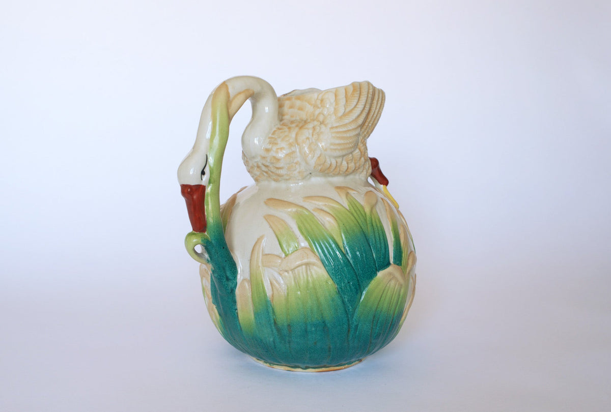 The Euro Trash Vintage Swan Motif Ceramic Pitcher features a white ceramic swan shape with a graceful swan handle and hand-painted green and yellow cattail designs at the base, set against a plain white background.