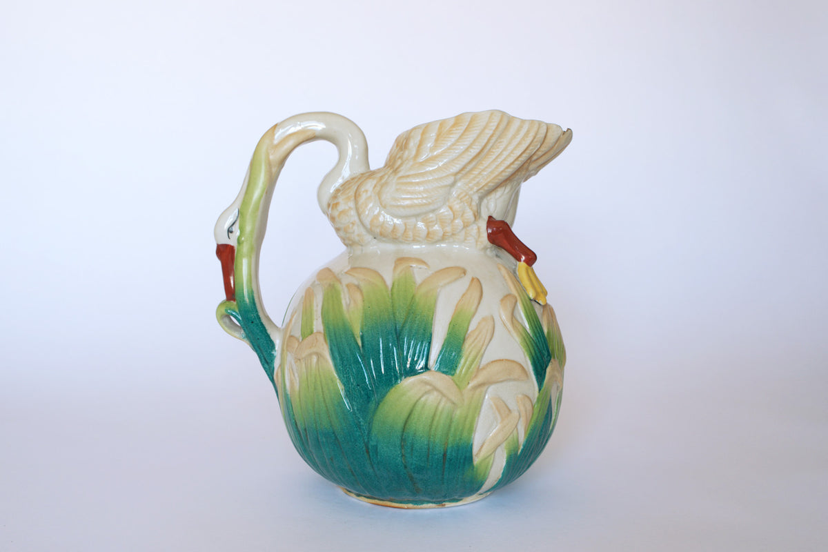 The Euro Trash Vintage Swan Motif Ceramic Pitcher features a swan-shaped handle, hand-painted with green and white cattail patterns and a red beak, displayed on a plain white background.