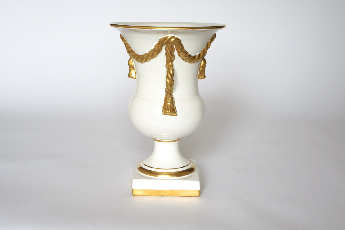The Euro Trash Vintage White & Gold Urn Vase features a neoclassical design with a draped garland detail, gold accents, and stands on a square gold-trimmed base against a light background.