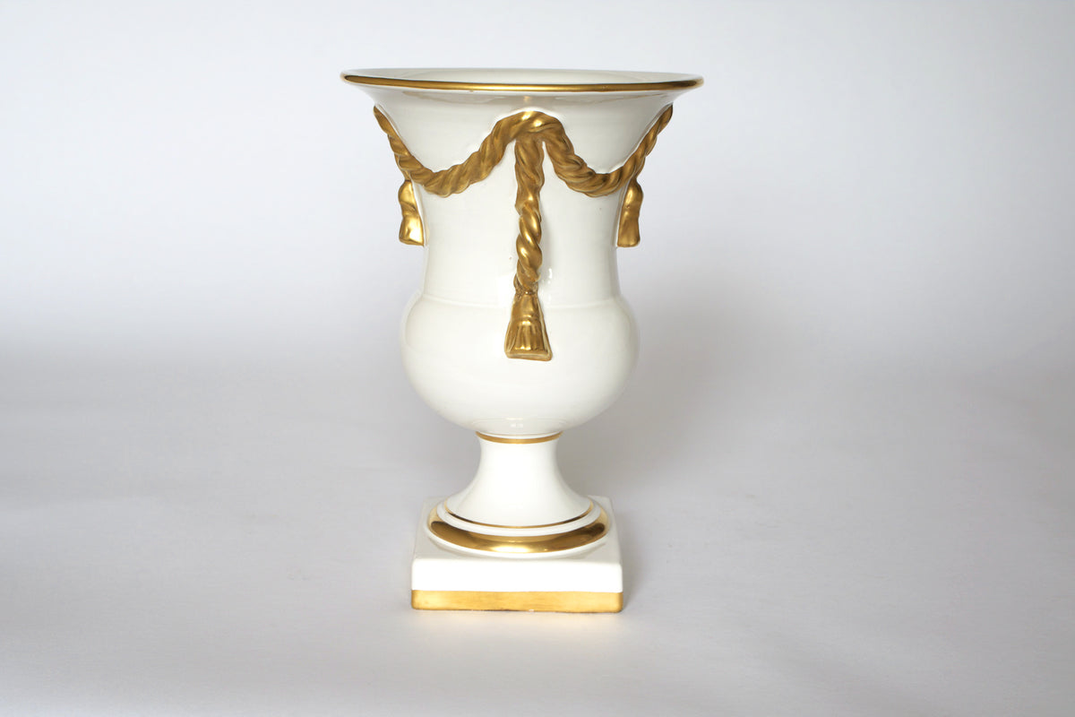 The Vintage White & Gold Urn Vase with Draped Garland Detail by Euro Trash features gold garland and tassels on white ceramic with a gold-trimmed square base, adding timeless elegance to any décor.