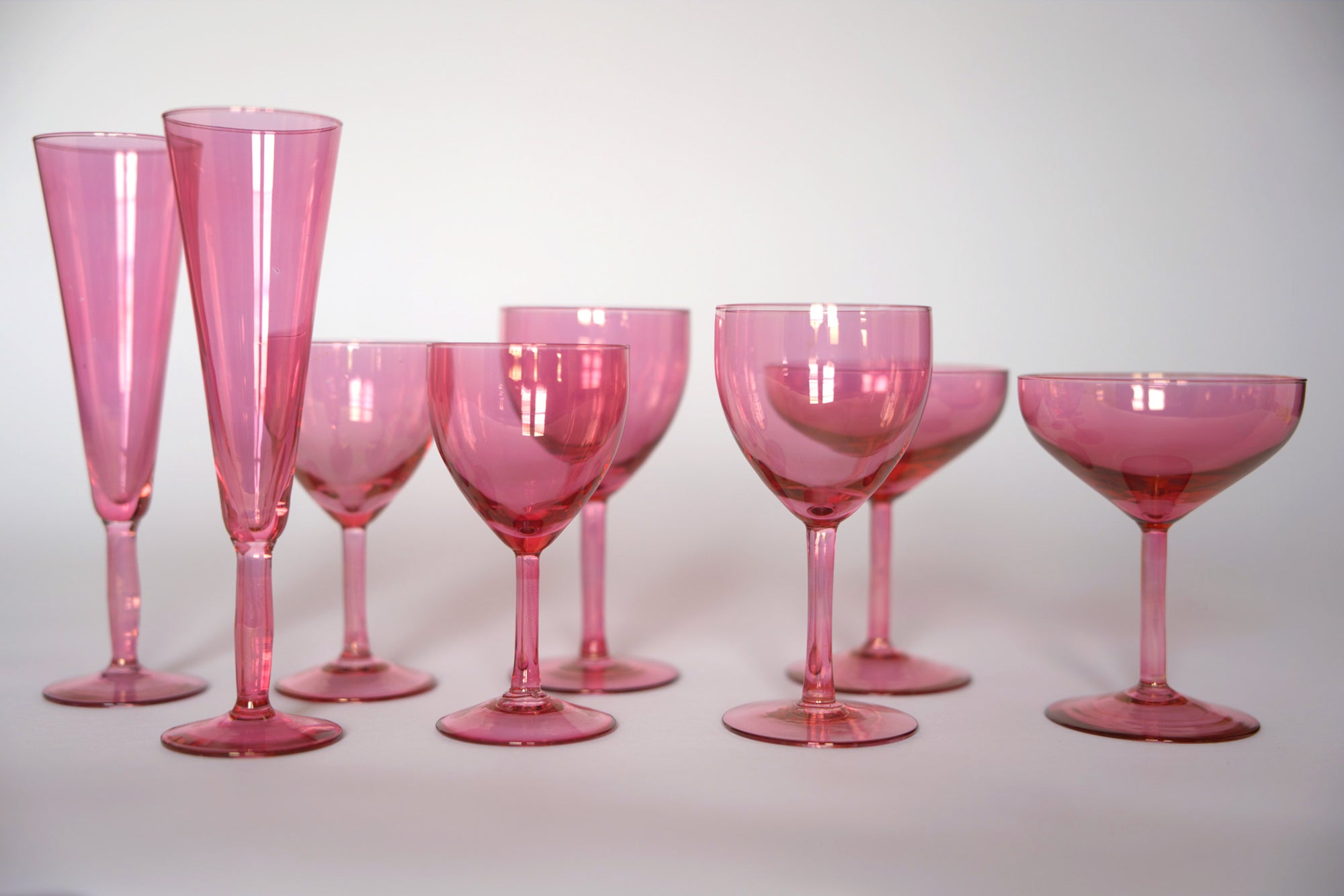 The Euro Trash Set of Vintage Pink Glass Stemware features eight elegant pieces, including wine, champagne, and cocktail glasses with pink champagne flutes, displayed in rows on a white background.