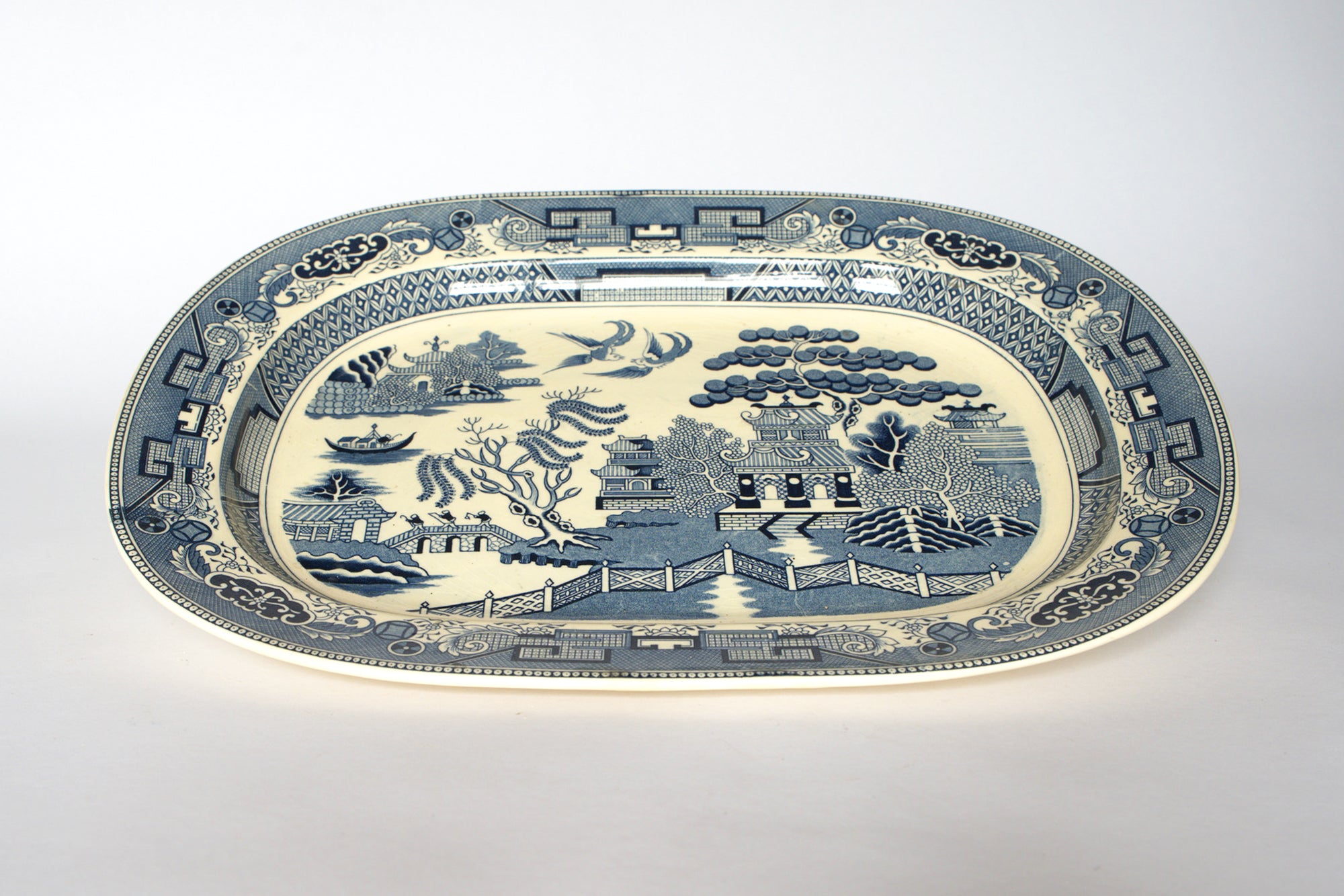 The Euro Trash Vintage Blue Willow Oval Serving Platter features a classic willow pattern with a pagoda, trees, bridge figures, birds, and a decorative border on a plain white background for timeless chinoiserie style.