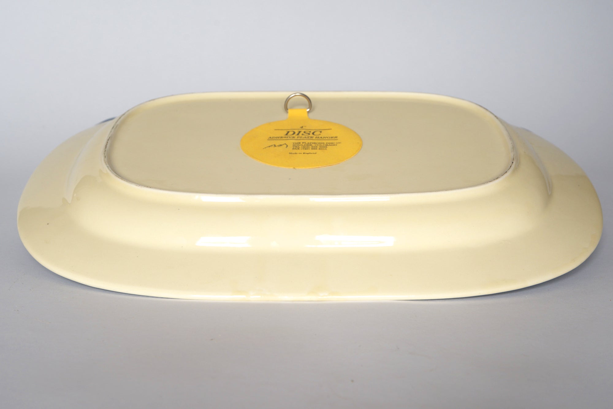 A rectangular, cream-colored Vintage Blue Willow Oval Serving Platter by Euro Trash is shown upside down, featuring a yellow circular label and metal hanging hook on its underside. Ideal as vintage chinoiserie decor, set against a plain light gray background.