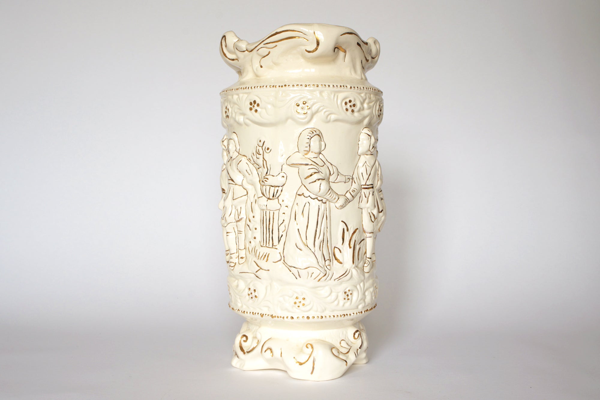The Euro Trash Vintage Figural Porcelain Vase is a cream-colored decorative piece with embossed scenes of people in traditional attire, featuring ornate details on the rim and base.