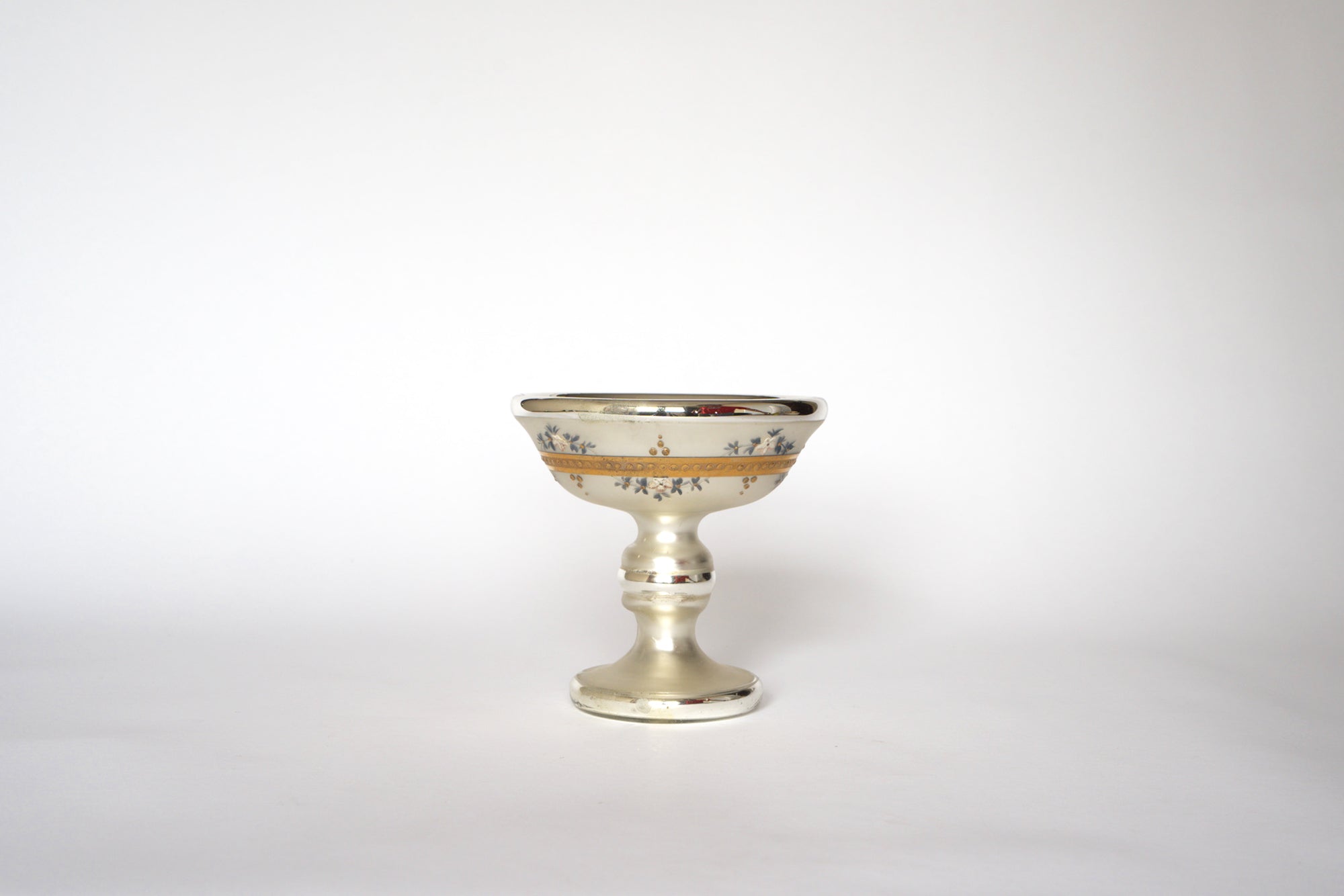 The Euro Trash Vintage Hand-Painted Mercury Glass Compote features delicate floral designs on ornate silver and gold glass, with a wide bowl and short pedestal base, displayed against a plain white background.