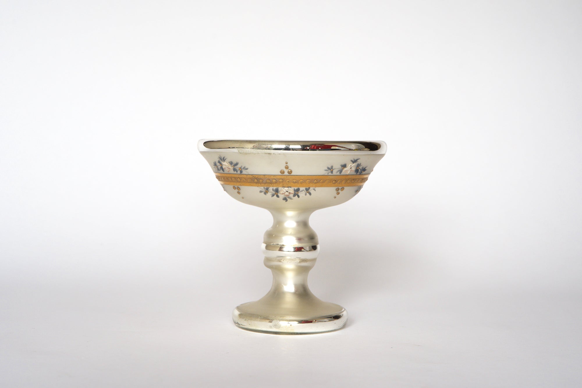 The Euro Trash Vintage Hand-Painted Mercury Glass Compote features a gold rim, floral details, and decorative bands on a short ornate stem, displayed against a plain white background.