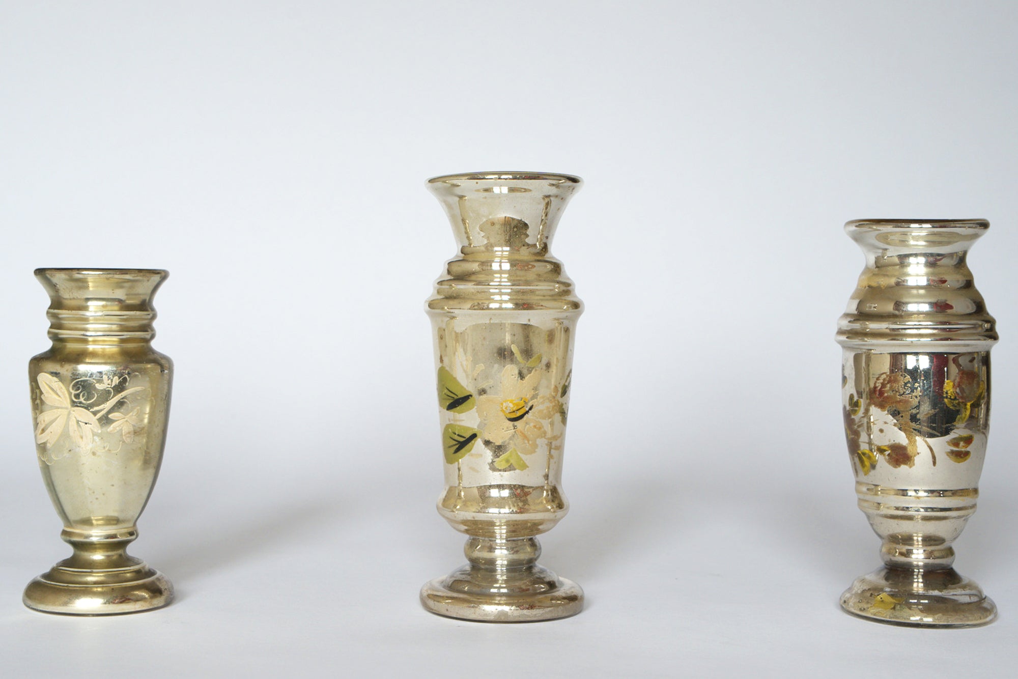 The Euro Trash Vintage Hand-Painted Mercury Glass Vase Set features three silvered vases with faded floral patterns, each uniquely shaped and showing charming signs of age, displayed against a plain white background.