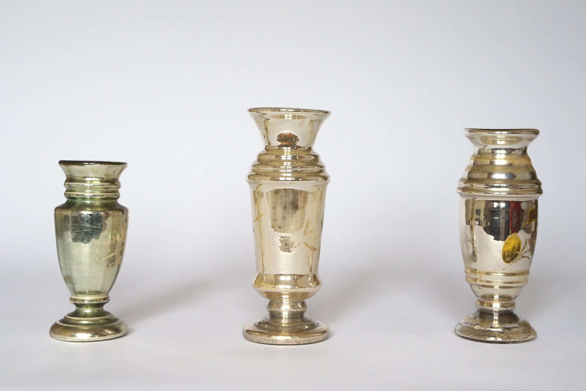 The Euro Trash Vintage Hand-Painted Mercury Glass Vase Set includes three vases of different shapes and heights, each with a reflective, gently tarnished finish for timeless appeal, displayed against a plain white background.