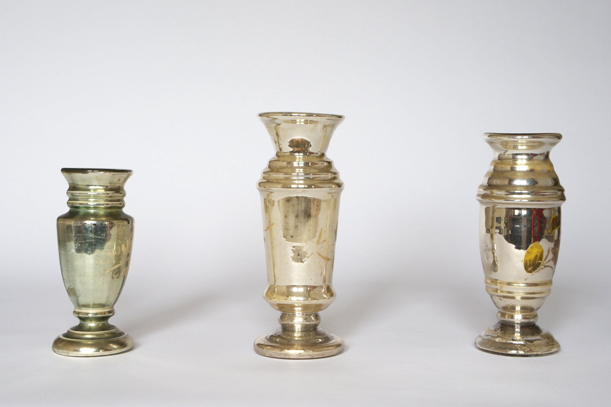 The Euro Trash Vintage Hand-Painted Mercury Glass Vase Set features three uniquely shaped vases with reflective, silvered finishes and charming tarnish detail, creating an elegant, antique display against a plain white background.