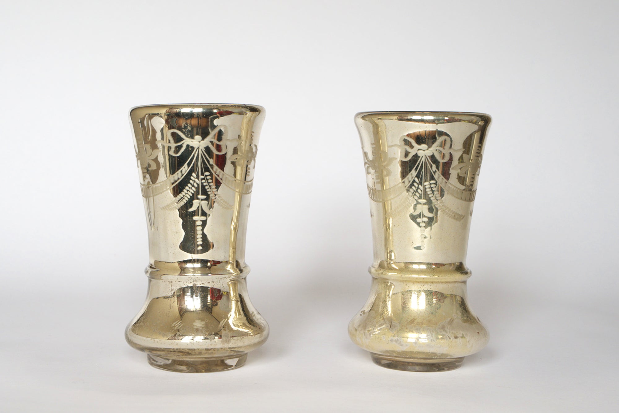 The Pair of Vintage Etched Mercury Glass Vases by Euro Trash features two silver-toned vases with etched garland motifs and flared tops, shown side by side against a plain white background.