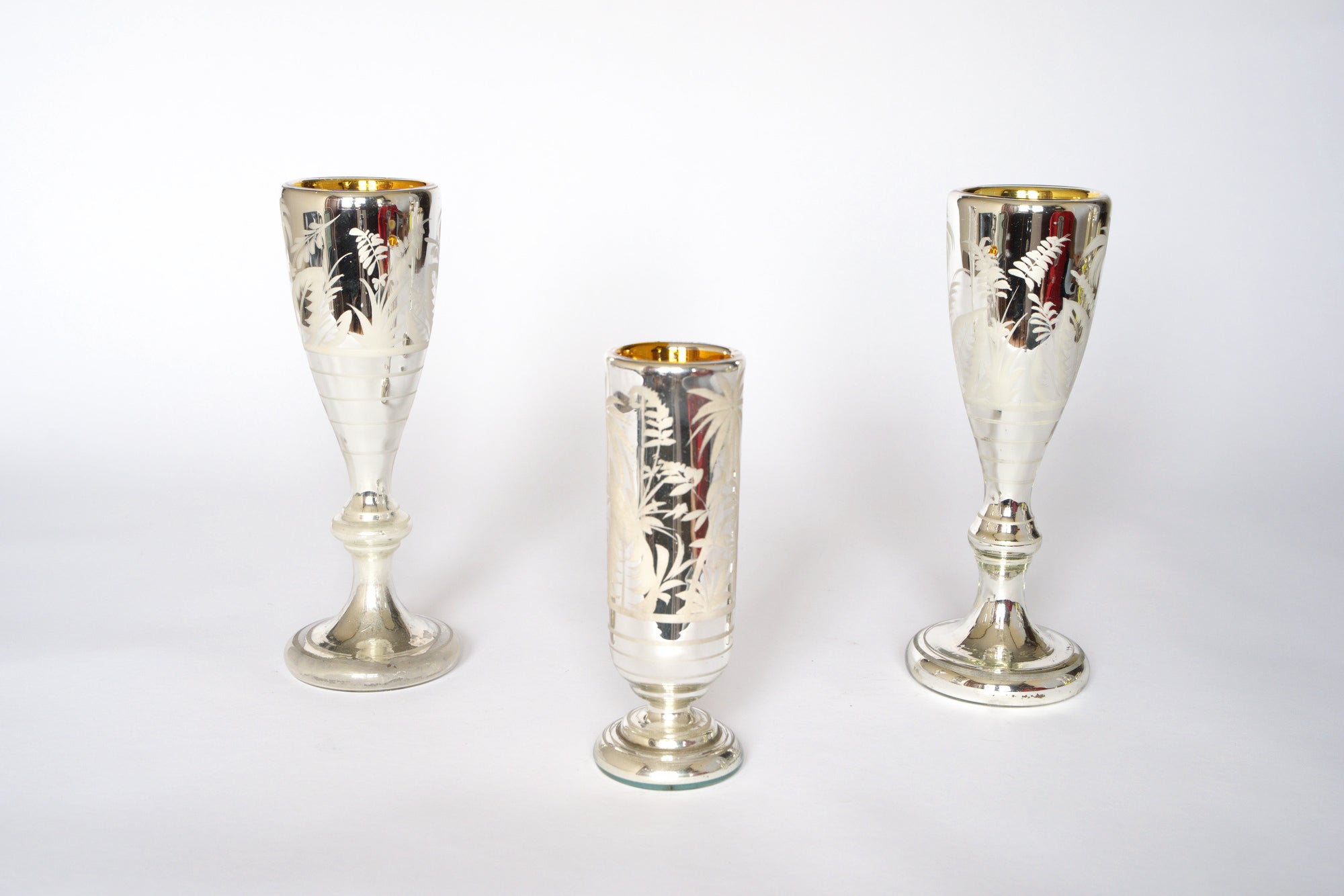 The Vintage Etched Mercury Glass Vessel Set by Euro Trash features three silvered vessels with frosted botanical patterns and gold-toned interiors. Two taller pieces frame the shorter center one, shown on a white background.